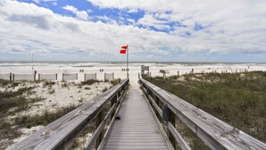 Una pasarela de madera que conduce a una playa con una bandera roja, dunas arenosas y un cielo nublado.