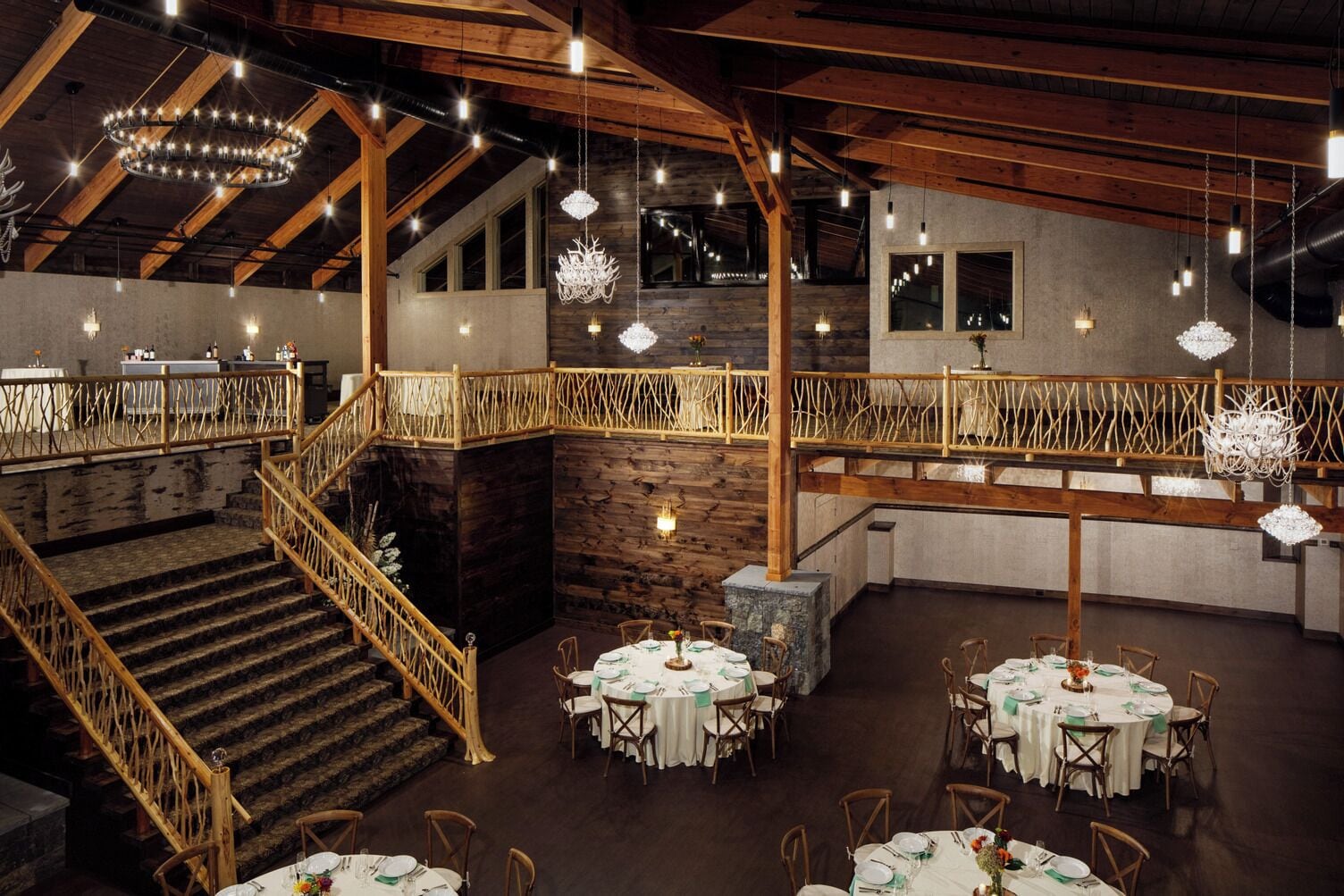 A spacious dining area with multiple round tables set for guests, a wooden staircase, and a high ceiling with exposed beams and hanging lights.