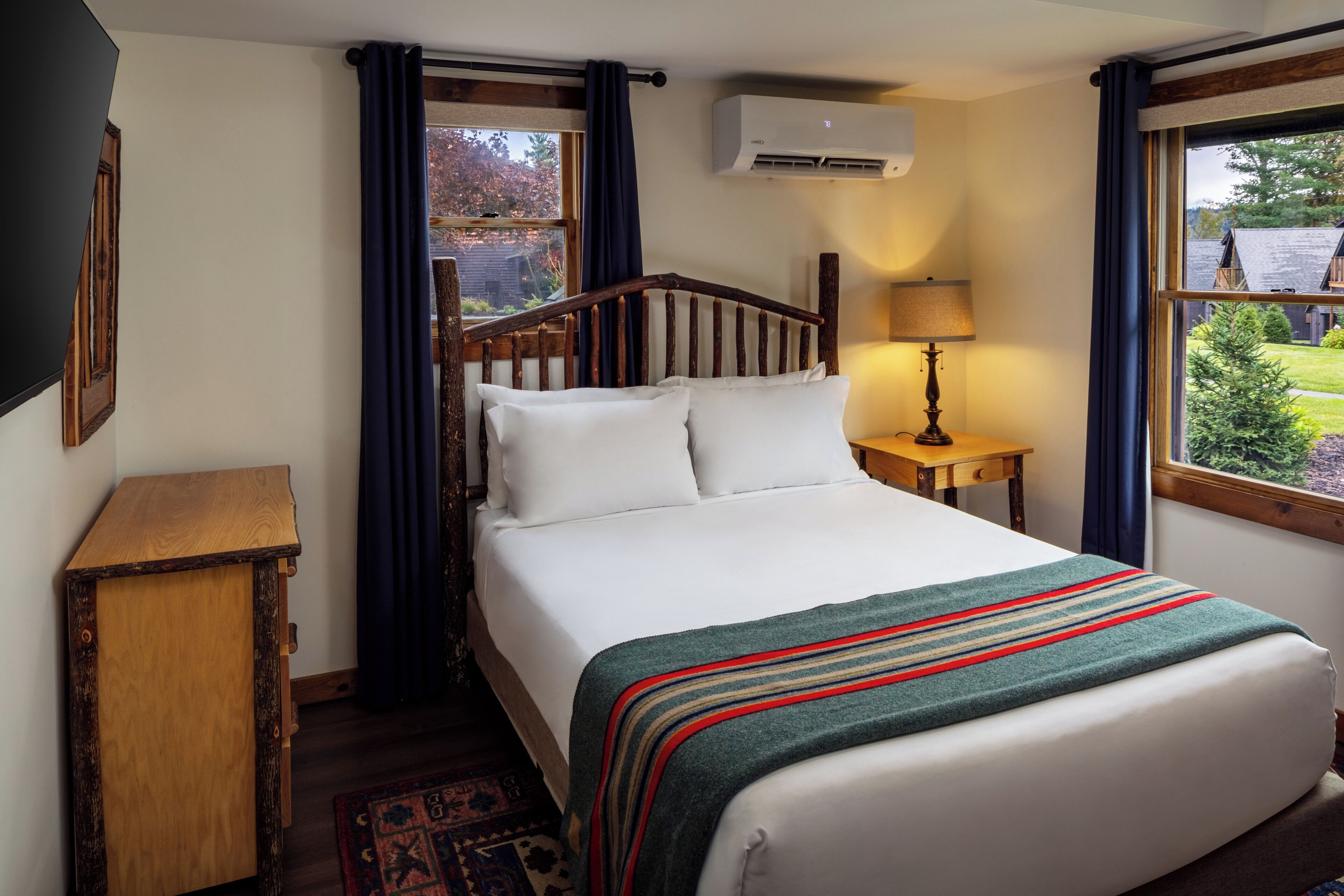 Un dormitorio con cabecera de madera, cama con ropa blanca, mesita de noche con lámpara y vistas al exterior a través de una ventana.
