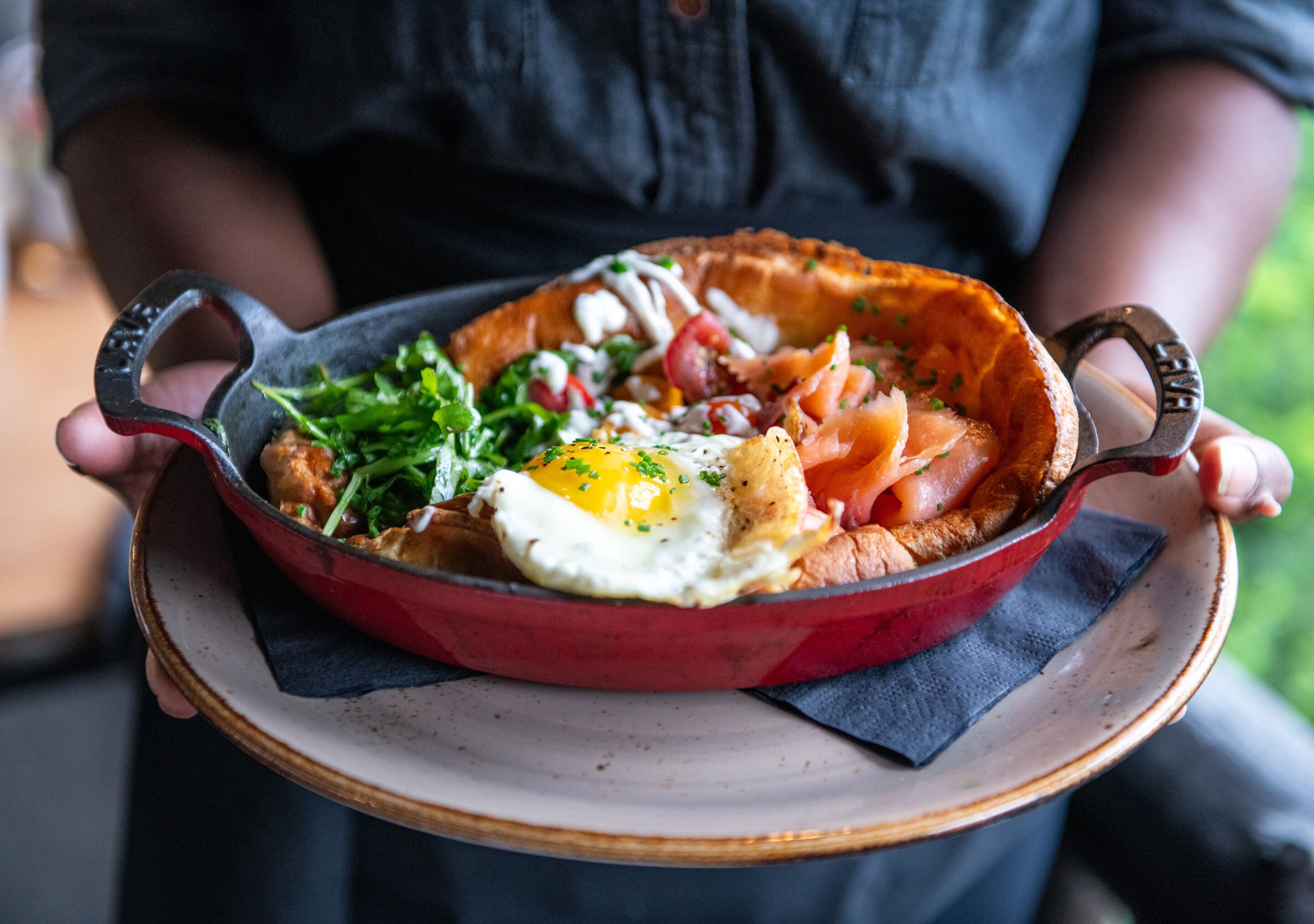 A person holding a red skillet with a breakfast dish containing eggs, greens, and meat.