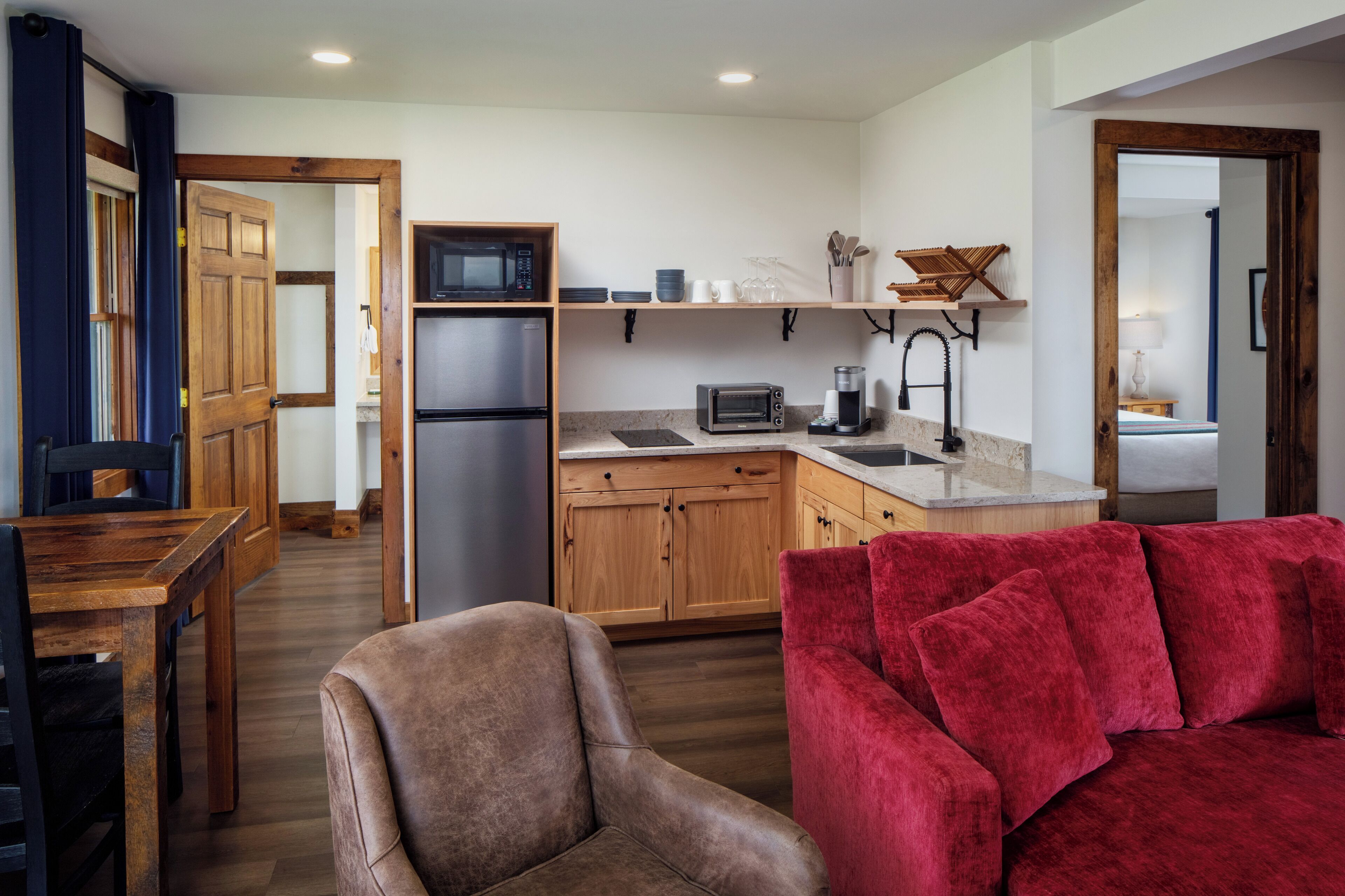 A compact kitchen with wooden cabinets, a refrigerator, microwave, and a toaster. There is a red sofa and a brown armchair in the living area.