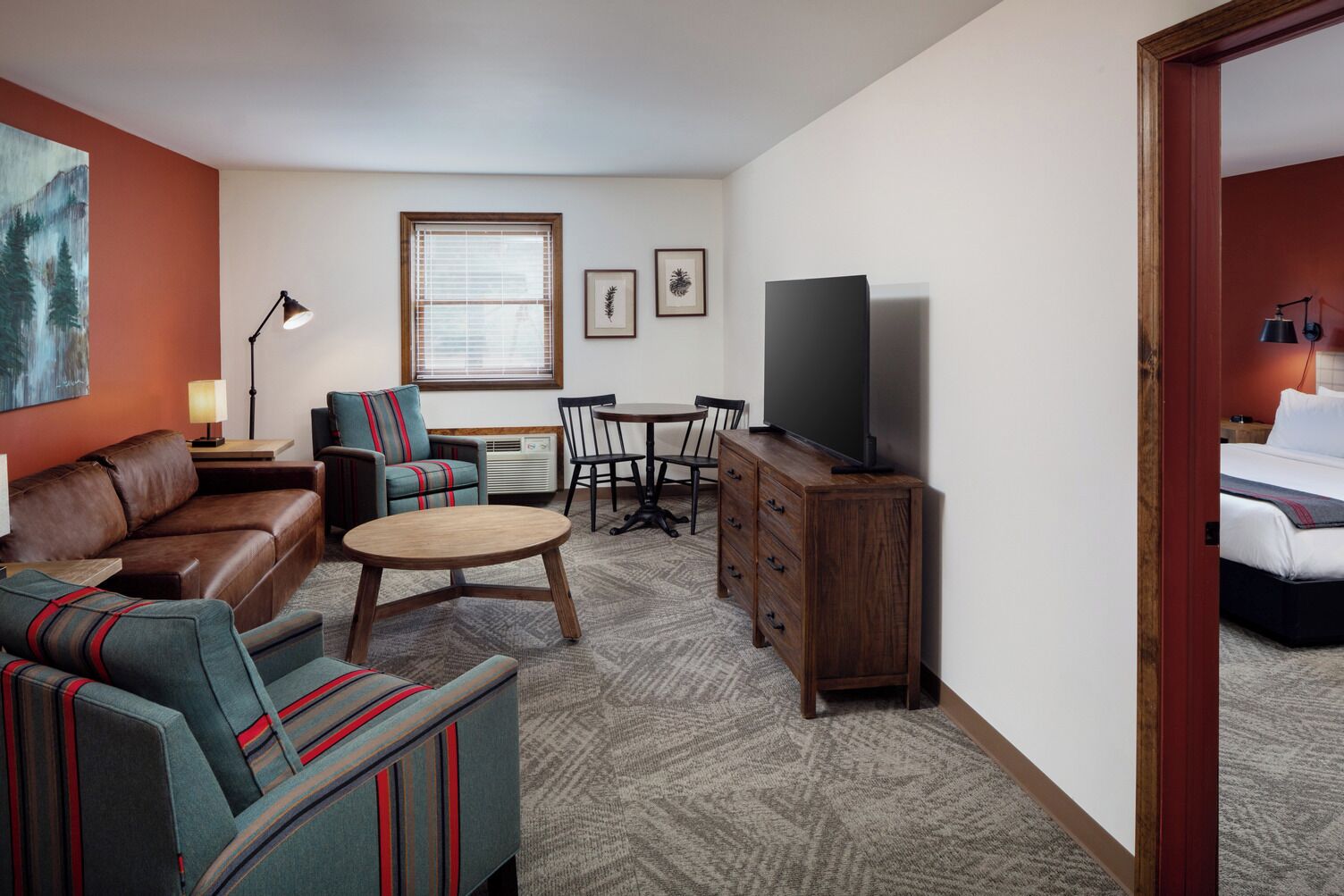 Une chambre d’hôtel avec un canapé en cuir marron, des fauteuils à rayures, une table basse en bois, un téléviseur posé sur un meuble en bois, une table à manger avec des chaises et une fenêtre avec des stores.