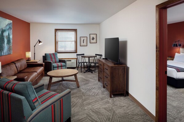 Una habitación de hotel con sofá de cuero marrón, sillones a rayas, una mesa de centro de madera, un televisor sobre un mueble de madera, una mesa de comedor con sillas y una ventana con persianas.
