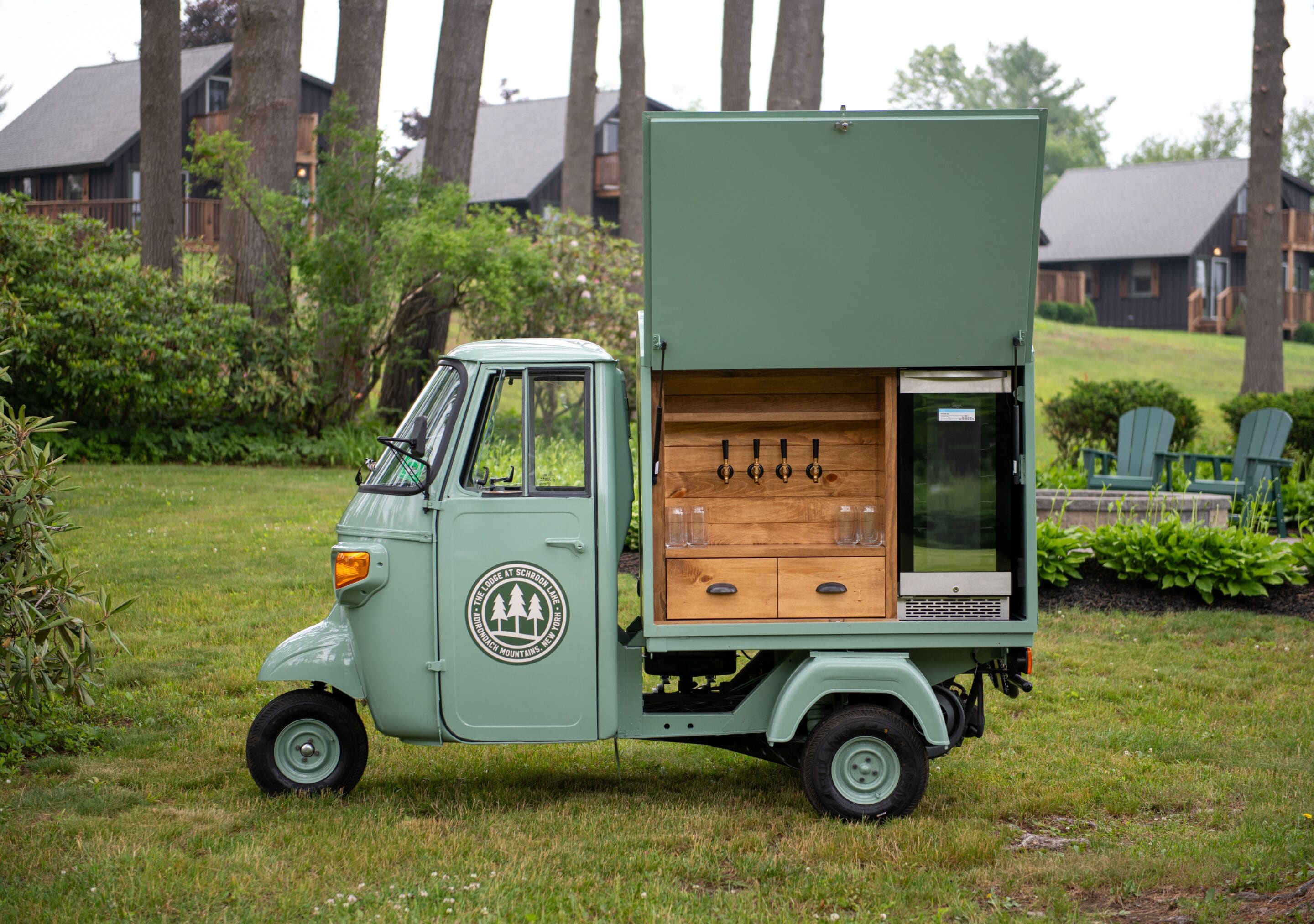A vintage green truck with a wooden storage box on the back, parked on grass with trees and a house in the background.