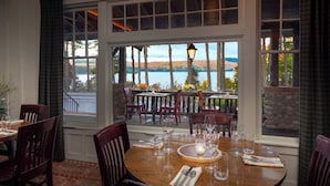 A dining area with a table set for four, large windows offering a view of a lake and trees, and a stone wall in the background.