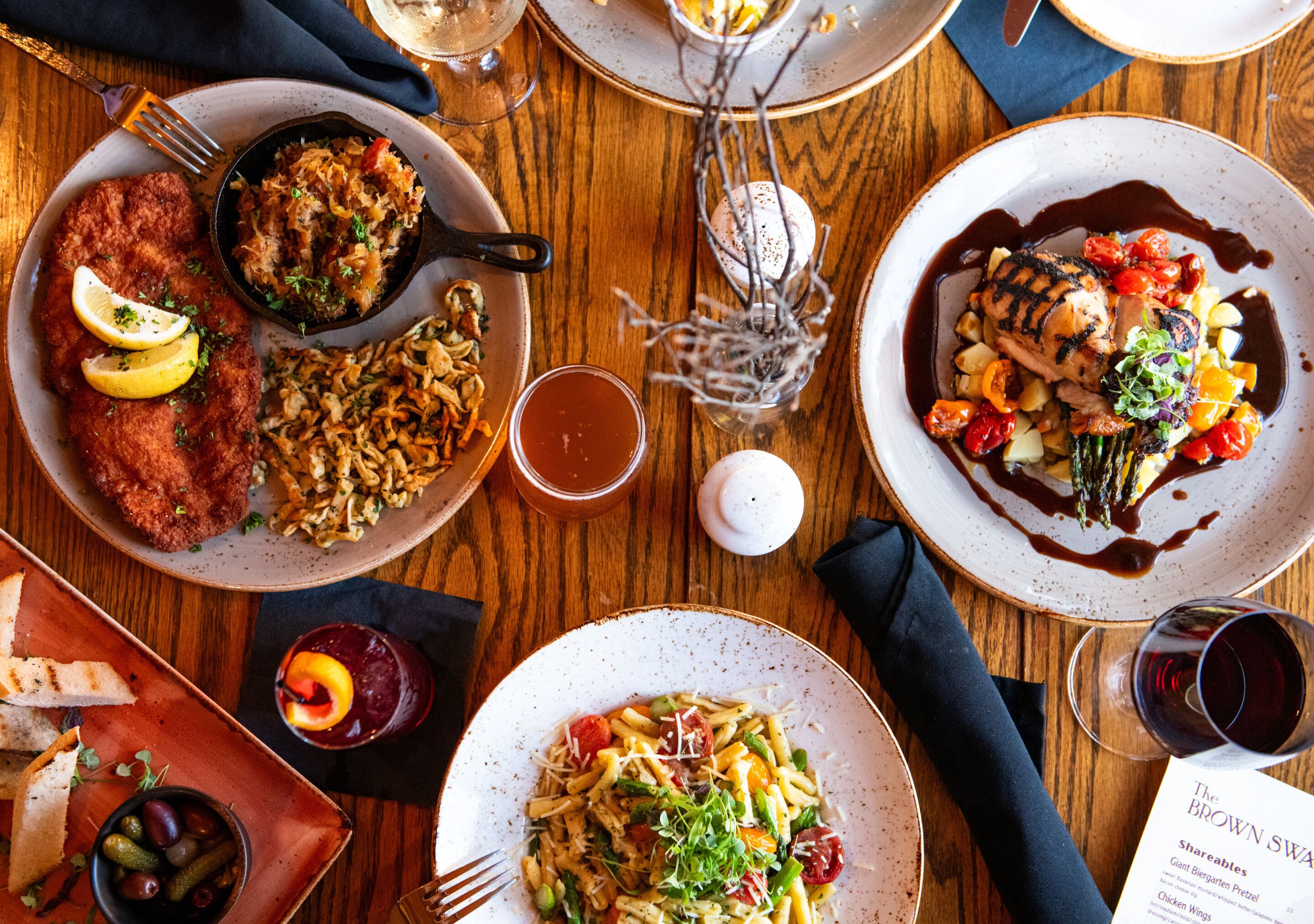A wooden table with various dishes, including a schnitzel, pasta, and grilled chicken, along with glasses of wine and a menu.
