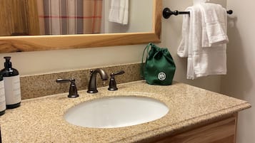 Un baño con encimera de granito, lavamanos blanco, tocador de madera, espejo y toallero con dos toallas blancas.