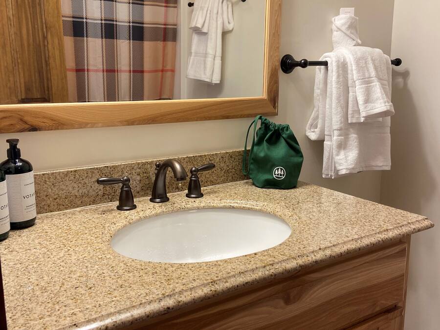 Un baño con encimera de granito, lavamanos blanco, tocador de madera, espejo y toallero con dos toallas blancas.