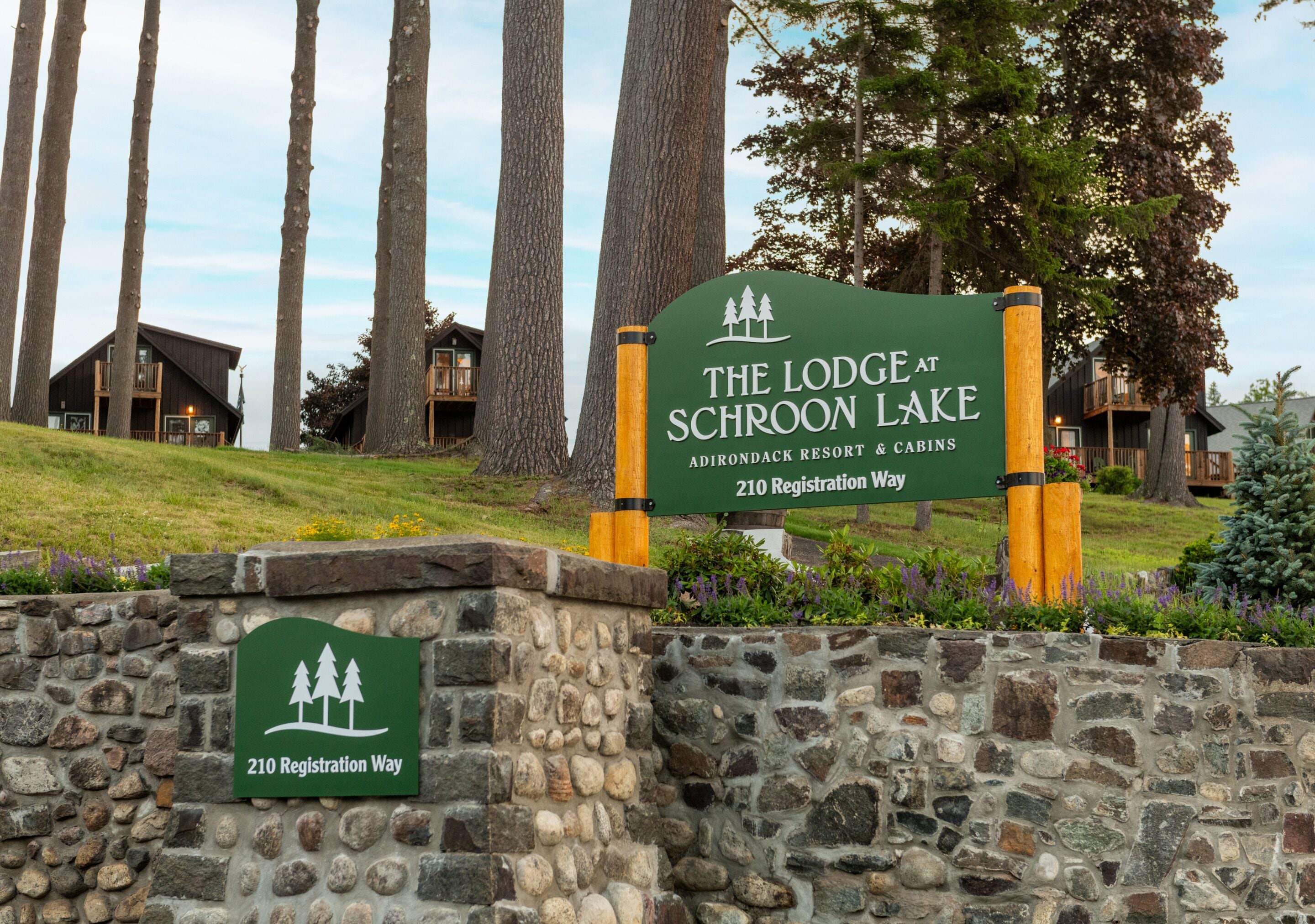 Señal de The Lodge at Schroon Lake, Adirondack Resort & Cabins, con un muro de piedra y árboles al fondo.