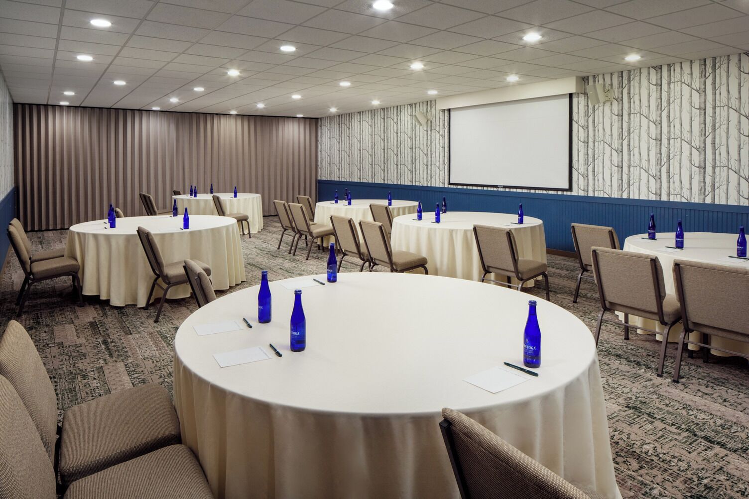 Un salón de conferencias con varias mesas redondas, cada una con sillas y botellas de agua azules.