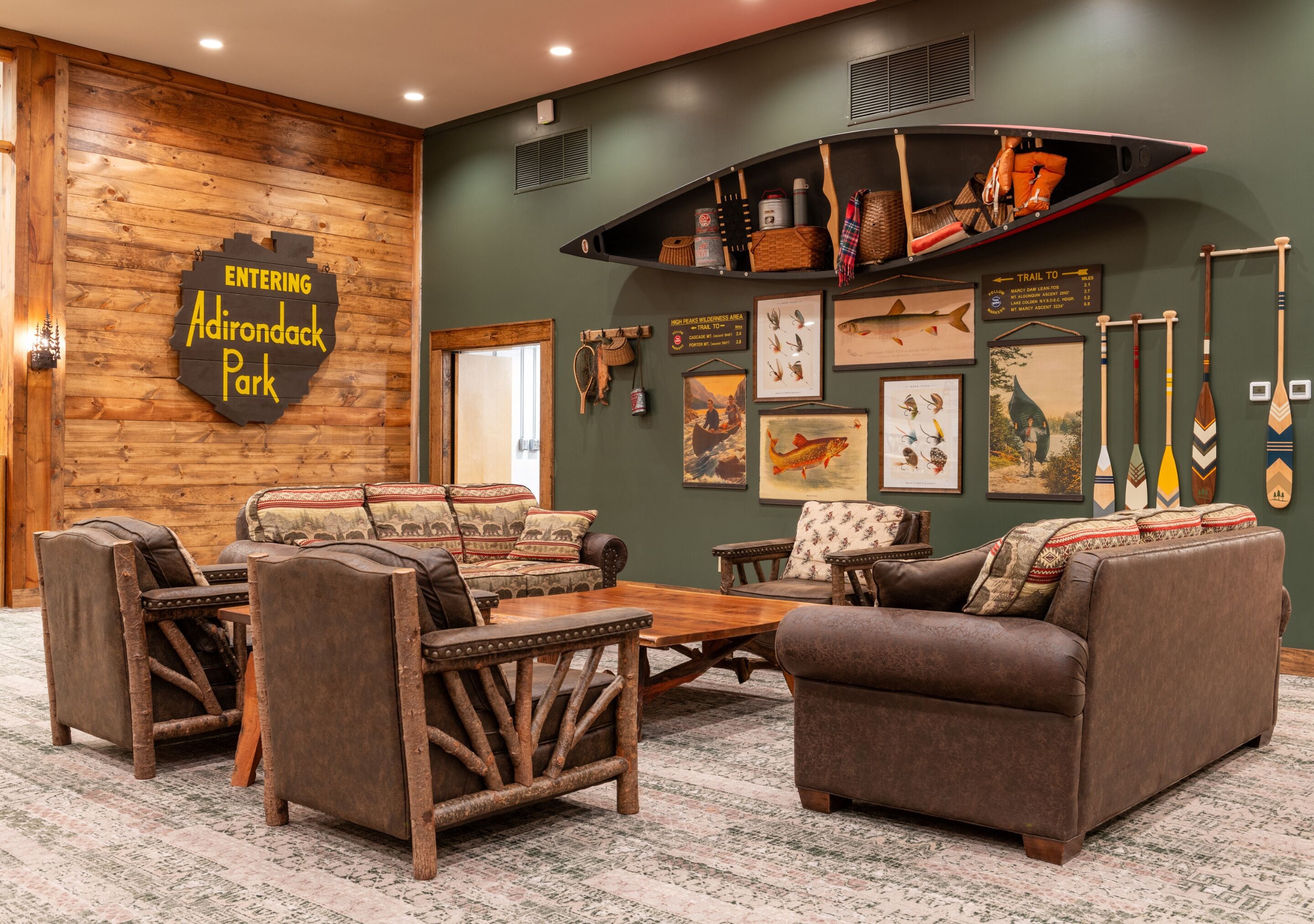 A cozy lounge area with rustic furniture, a wooden wall with a sign for Adirondack Park, and various fishing and paddling equipment displayed on the walls.