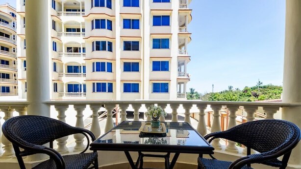 A balcony with a glass table and two wicker chairs, overlooking a multi-story building with balconies.