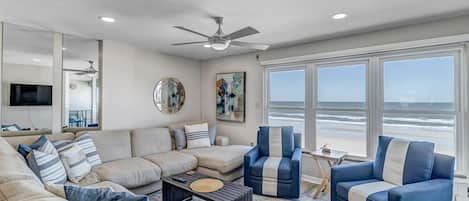 Uma sala de estar ampla com uma grande janela com vista para a praia, um ventilador de teto e um conjunto de sofás moderno.