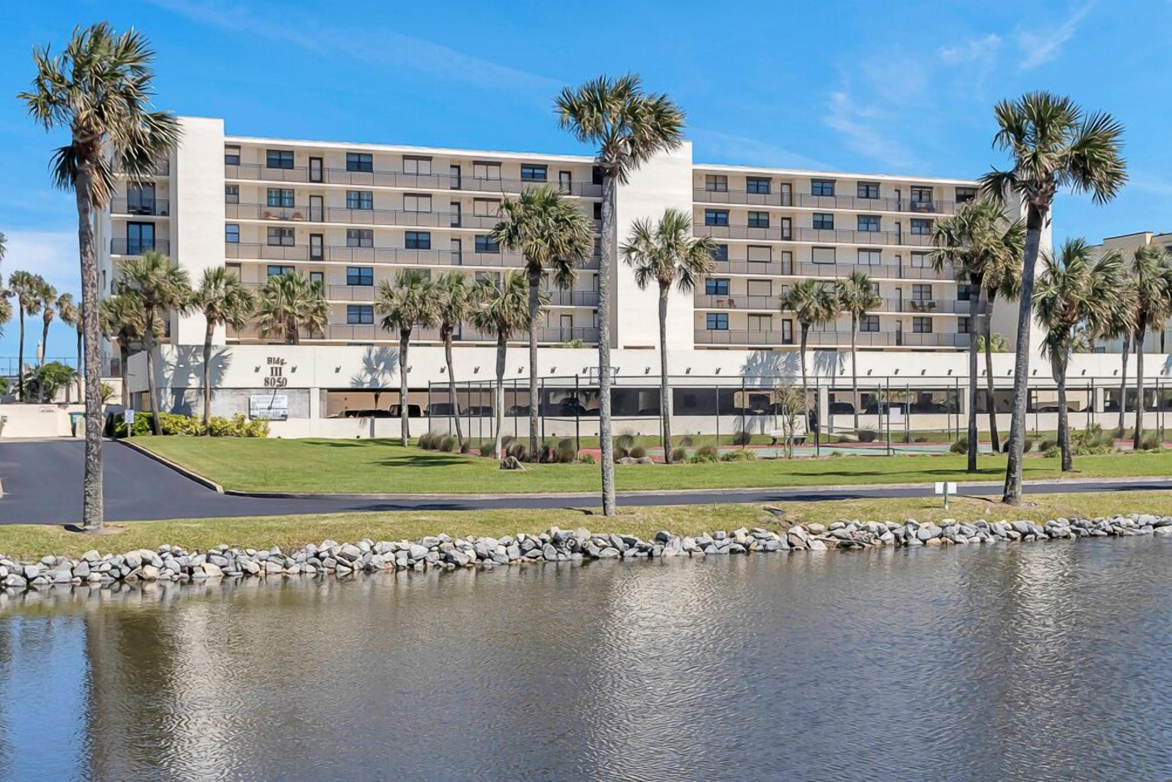 A hotel with a modern design, featuring a large building with multiple floors and balconies. In front of the hotel, there is a well-maintained lawn and a small pond with a stone border.