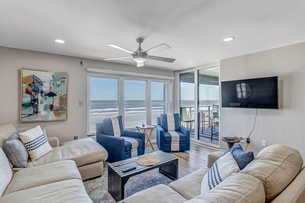 A spacious living room with a large window overlooking the ocean, a ceiling fan, and a flat-screen TV mounted on the wall.