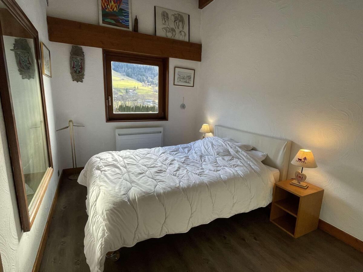 A bedroom with a bed, a nightstand with a lamp, a window with a view, and wall-mounted artwork.