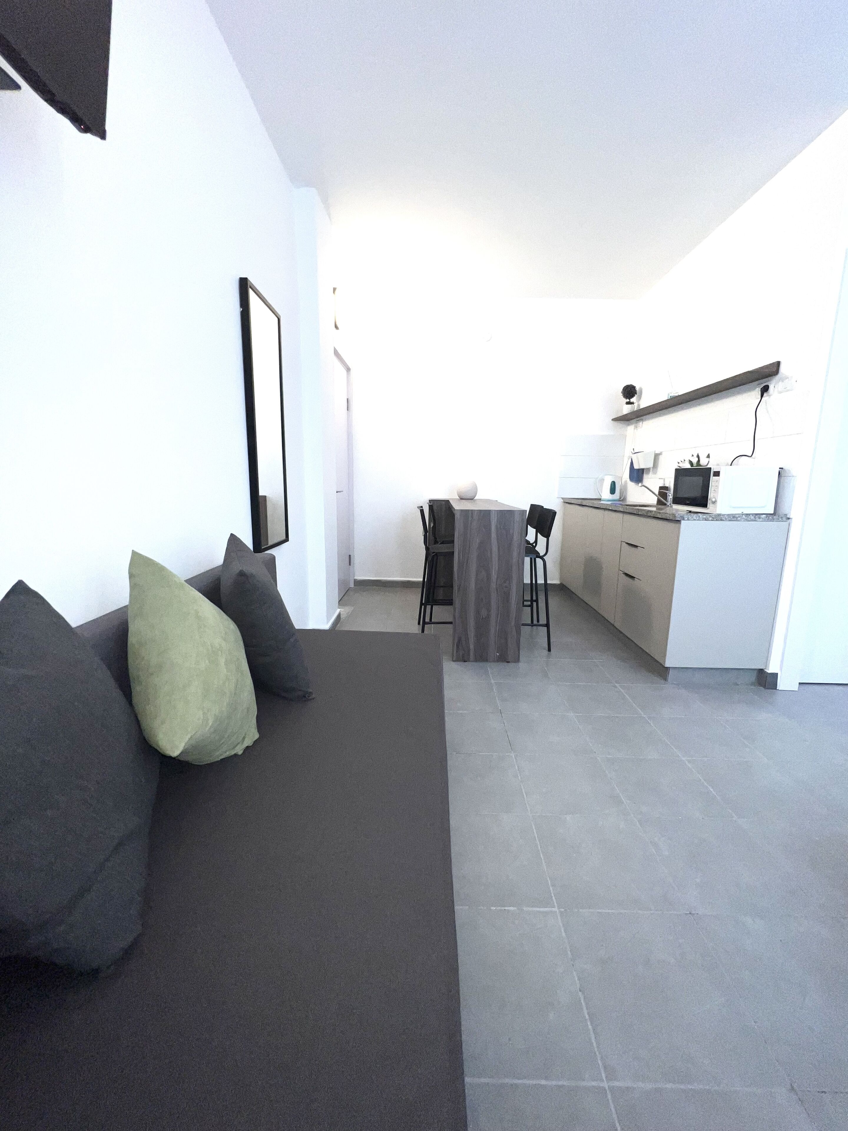 A modern living room with a dark sofa, a small dining table, and a kitchen area in the background.