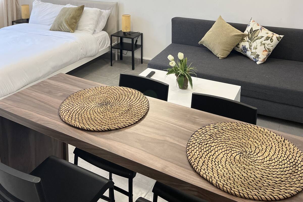 Una habitación moderna y bien iluminada con una cama, un sofá, una mesa de comedor con individuales tejidos y dos obras de arte colgadas en la pared.