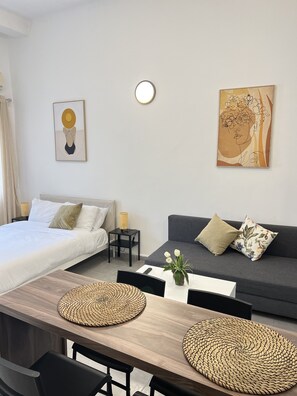 A modern, well-lit room with a bed, a sofa, a dining table with woven placemats, and two wall-mounted artworks.
