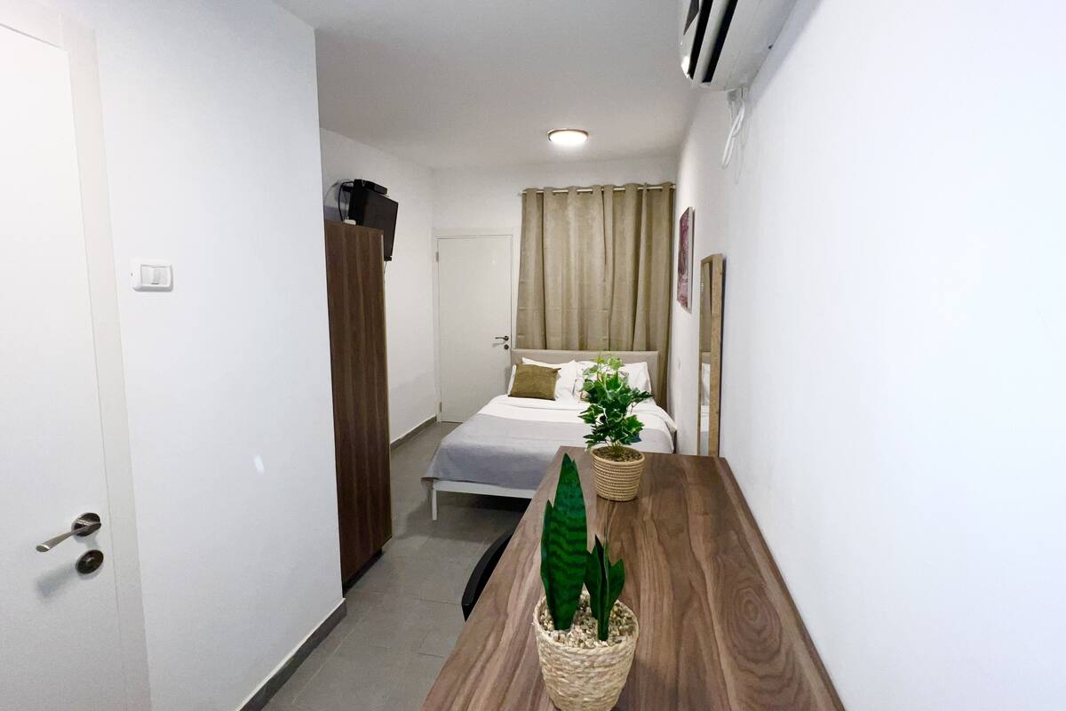 Una habitación de hotel con una cama, una mesita de noche de madera, una planta en maceta y un espejo.