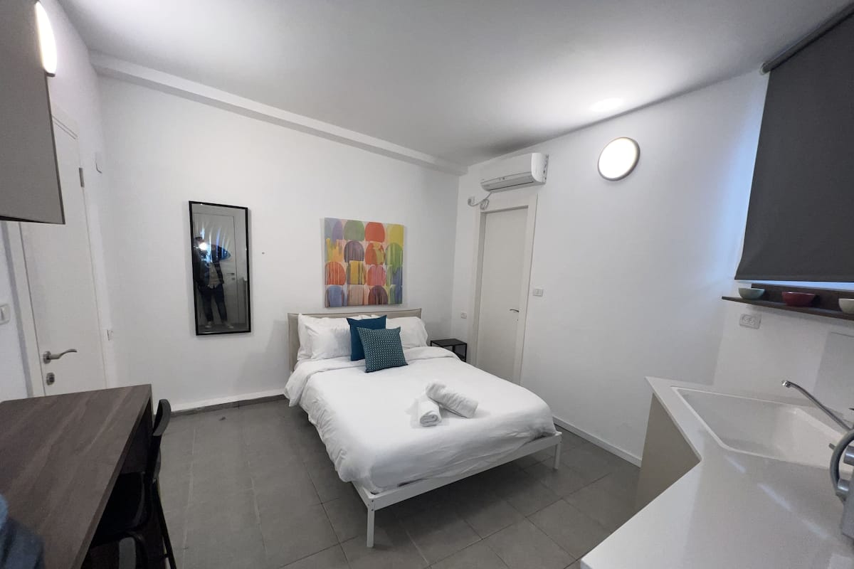 Una habitación de hotel moderna con una cama, un escritorio pequeño, una silla, un baño con lavabo y espejo, y un cuadro abstracto colorido en la pared.