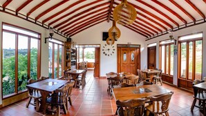 A spacious dining area with wooden tables and chairs, large windows offering a view of greenery, and a high wooden ceiling with exposed beams. - Hotel Cordillera de los Andes  (Villa de Leyva)