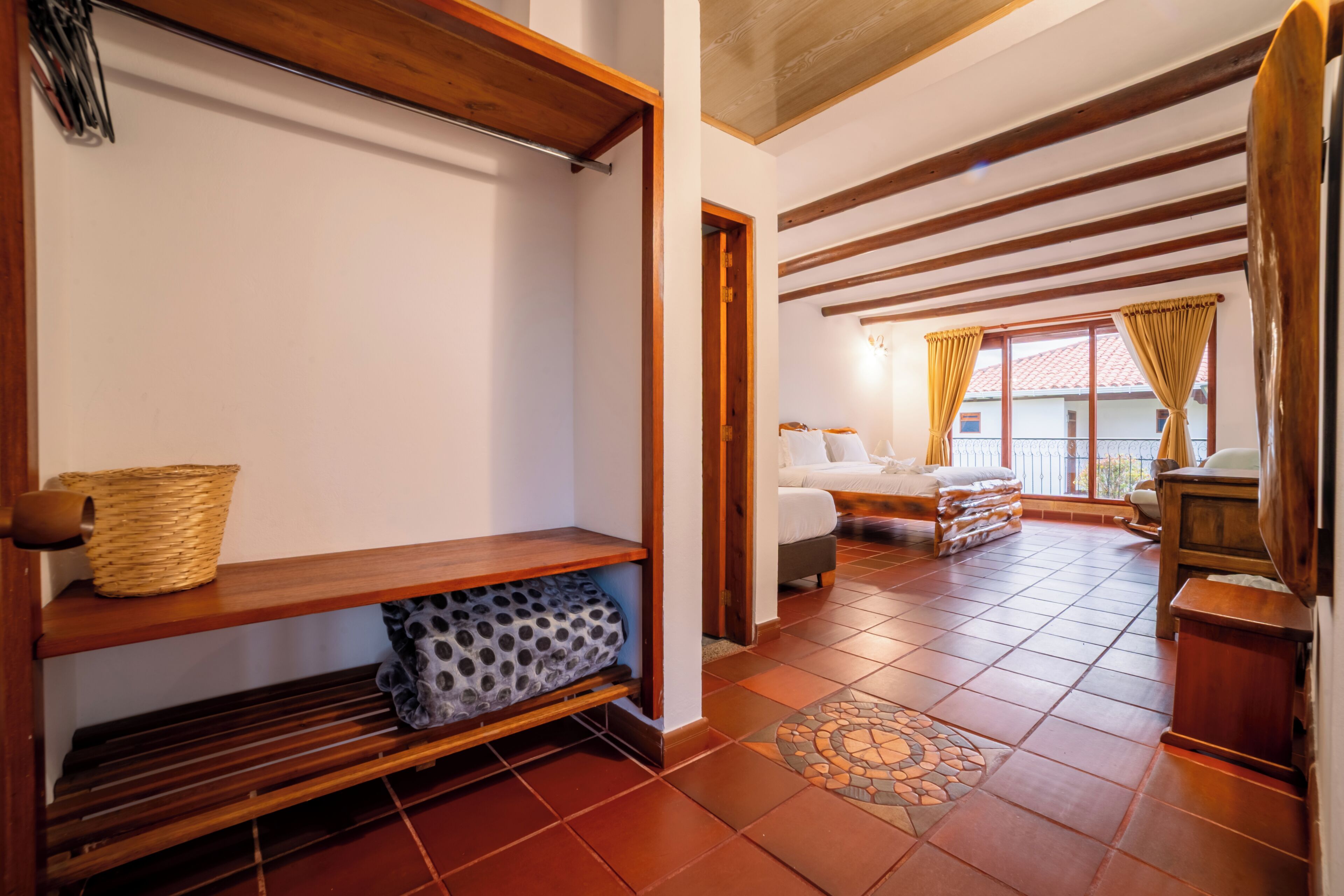 A room with a wooden bench, a patterned cushion, a woven basket, and a bed with a view through a window.