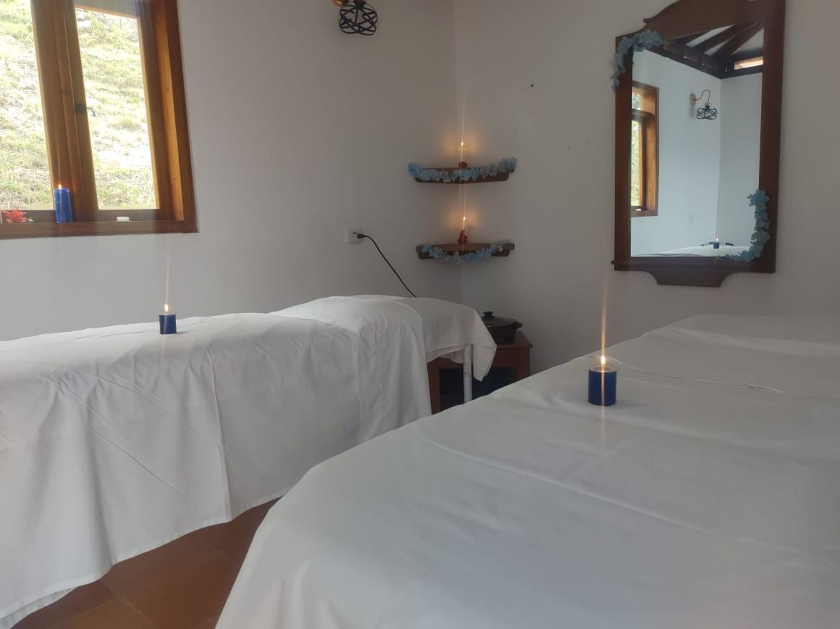 Una stanza con due letti, ognuno con una coperta bianca e una candela blu in un portacandela. Si vede una finestra a sinistra, uno specchio a destra e uno scaffale con candele e una piccola pianta.