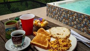 Ett frukostbord med rostat bröd, äggröra, en kopp kaffe och en skål med frukt, med utsikt över en pool och berg.