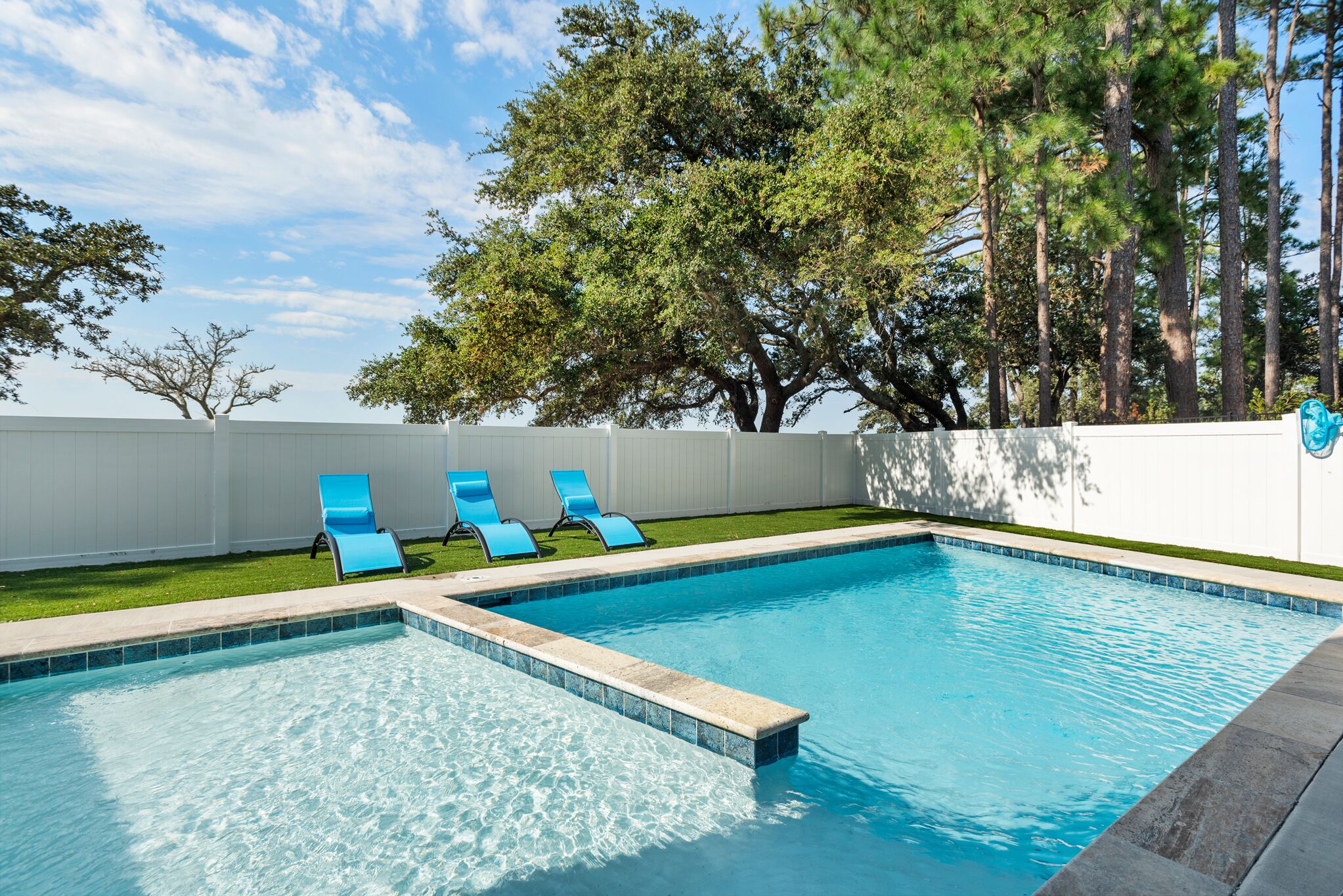 A rectangular swimming pool with blue tiles, surrounded by a white fence and trees.