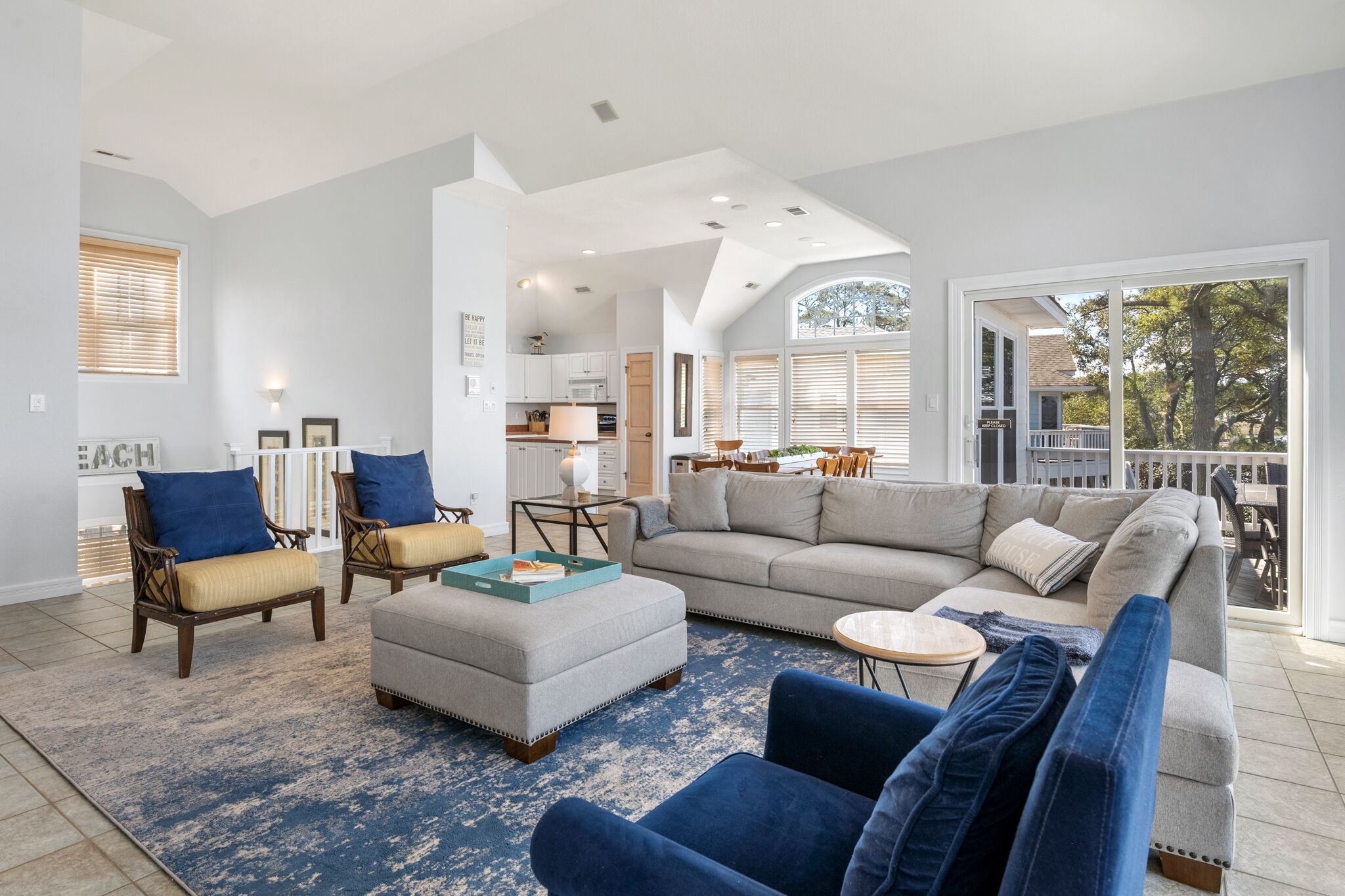 A spacious living room with a large gray sectional sofa, two armchairs, and a coffee table. A kitchen is visible in the background, and there is a balcony with a railing.