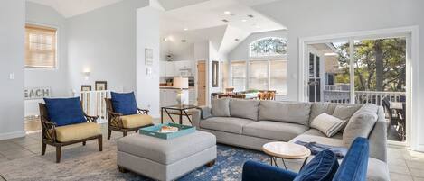A spacious living room with a large gray sectional sofa, two armchairs, and a coffee table. A kitchen is visible in the background, and there is a balcony with a railing.