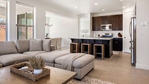 Una sala de estar moderna con un sofá modular, una mesa de centro con objetos decorativos y una cocina con armarios oscuros y electrodomésticos de acero inoxidable.