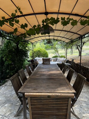 An outdoor covered patio with a wooden table and chairs, surrounded by greenery and a stone wall.