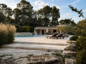 A pool area with lounge chairs, a stone wall, and a building with a tiled roof.