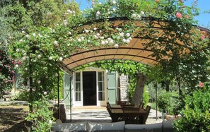 A covered outdoor seating area with a table and chairs, surrounded by lush greenery and flowering plants.