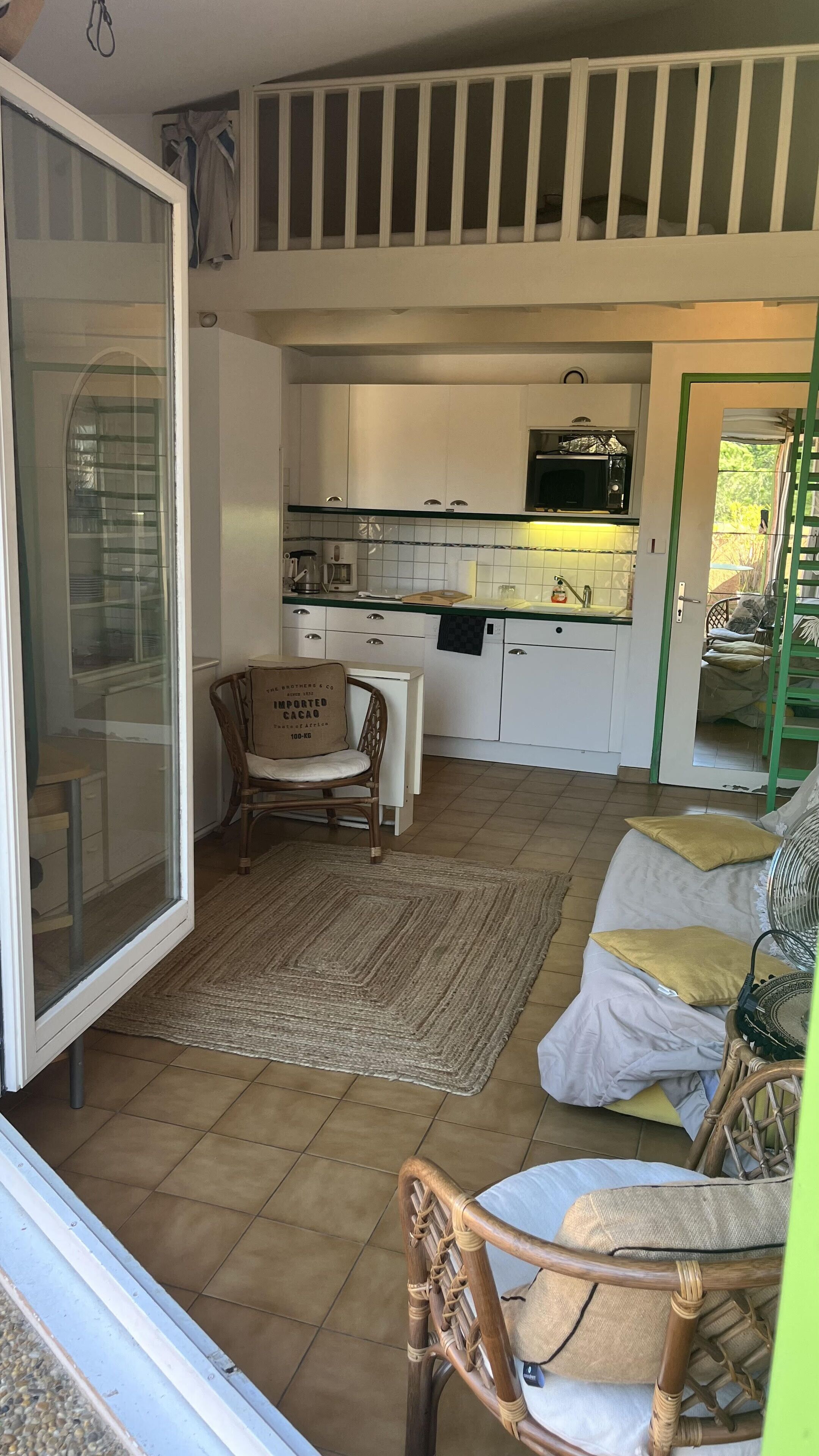 A room with a sliding glass door leading to a balcony, a wicker chair, a sofa with cushions, and a kitchen area with white cabinets and a microwave.