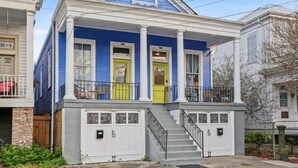 Une maison à deux étages, avec une façade bleue, des colonnes blanches, une porte d’entrée jaune et un porche avec une balustrade noire.