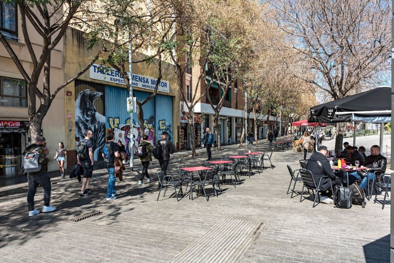 An outdoor café area with tables and chairs, a mural on the building, and people walking by.