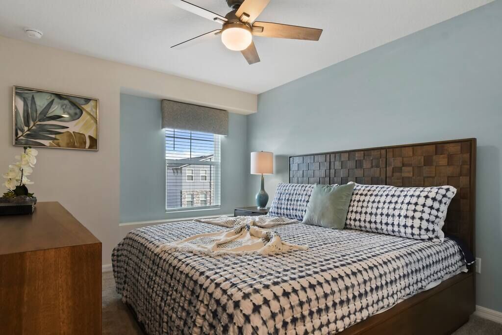 A bedroom with a bed, a ceiling fan, a window, and a bedside table with a lamp.