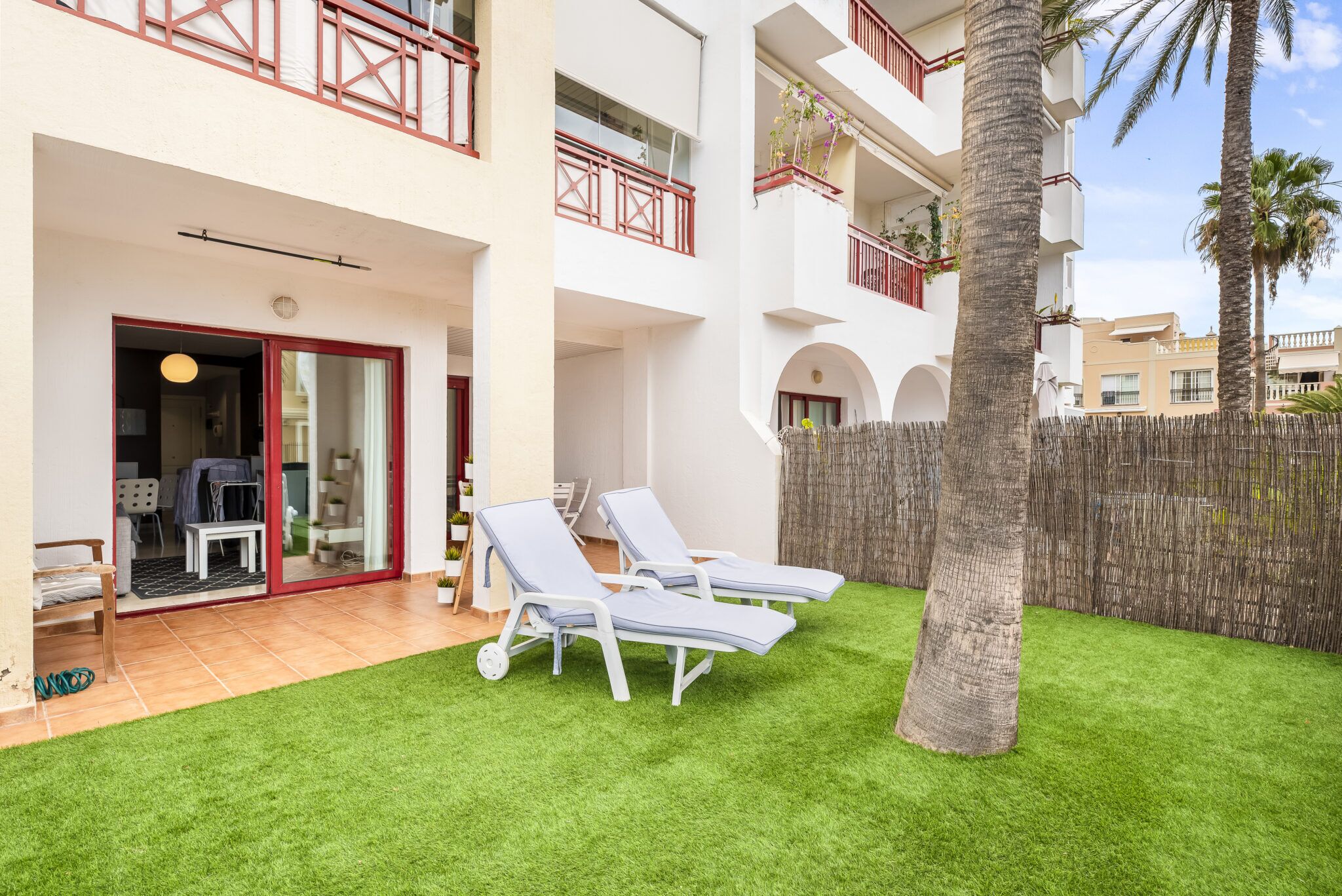 Een gebouw met twee verdiepingen, een balkon, een houten hek en twee loungestoelen op een groen gazon.