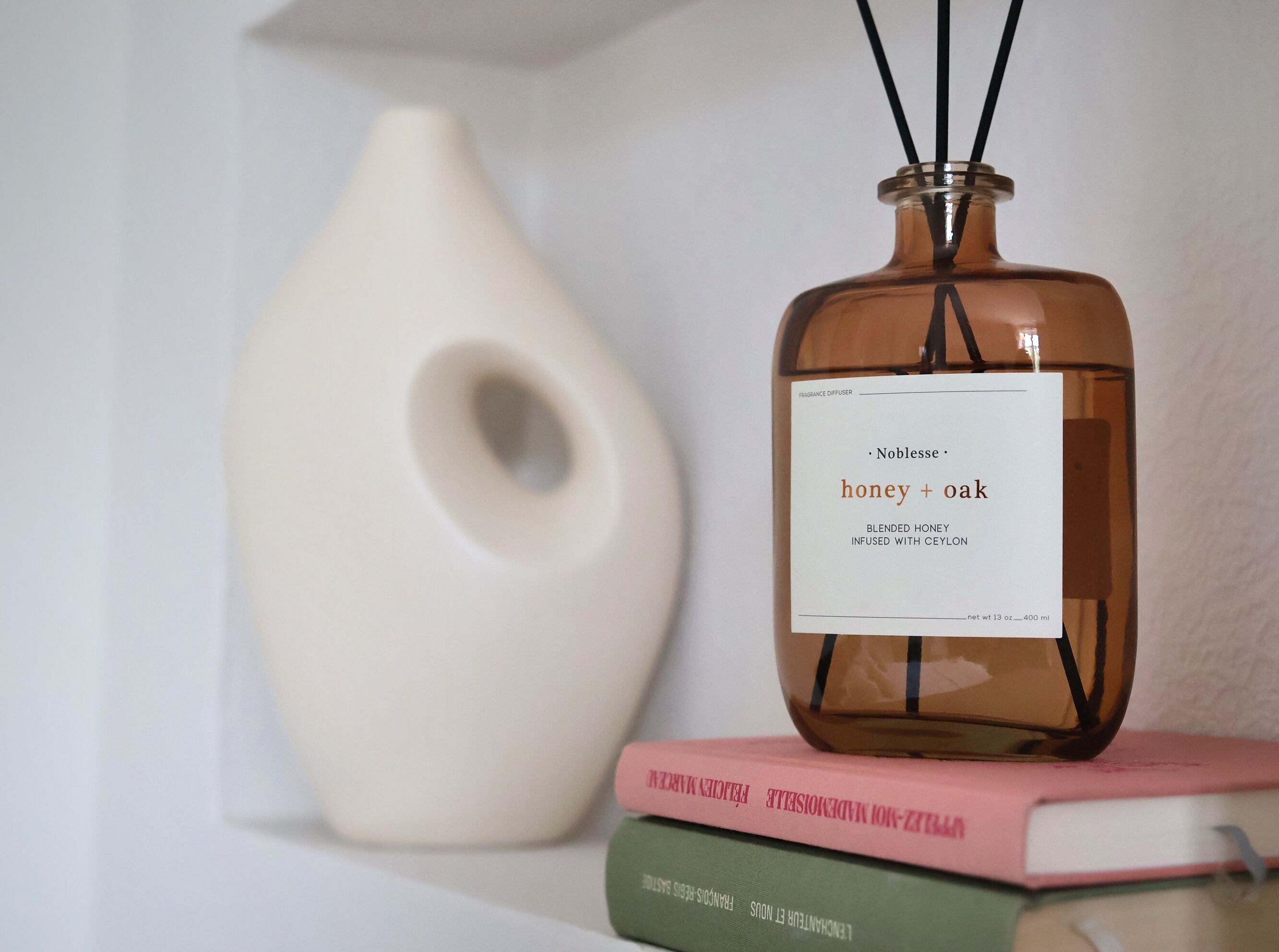 A bottle of fragrance diffuser with a label reading "Noblesse honey + oak" on top of a stack of books.