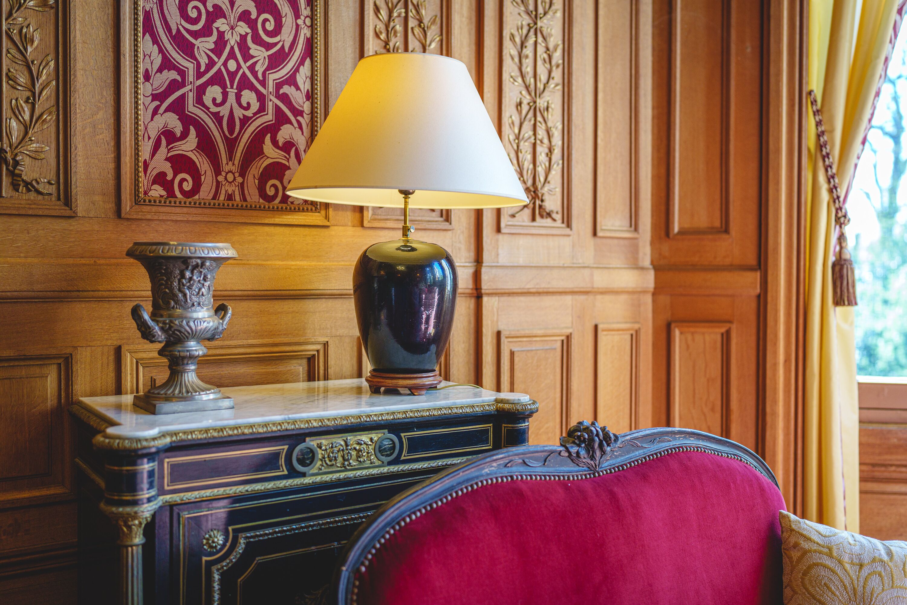 A room with a wooden paneled wall, a decorative lamp, a vase, and a red upholstered chair.
