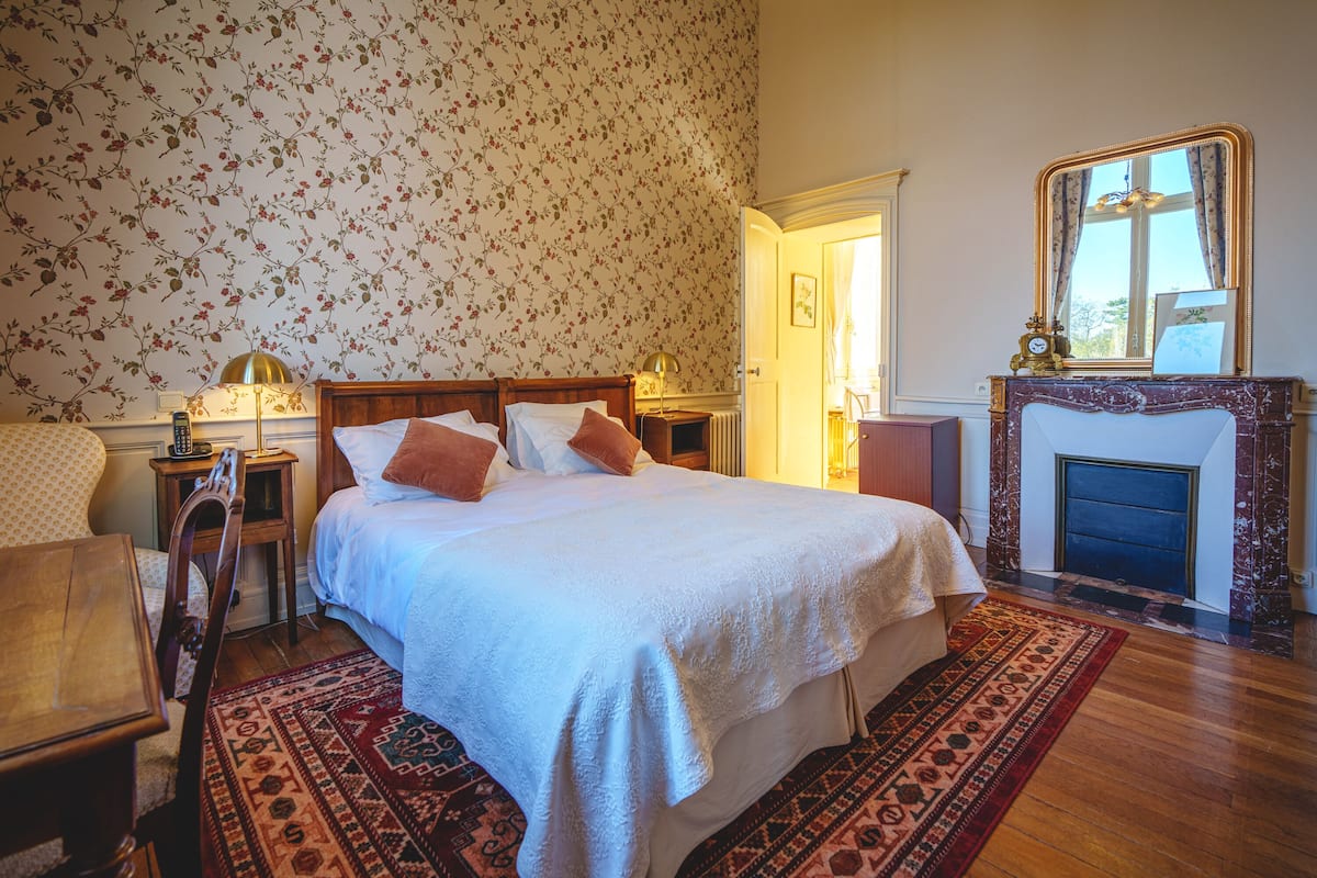 A bedroom with a large bed, a wooden nightstand, a bedside table with a lamp, a patterned rug, and a fireplace with a mirror above it.