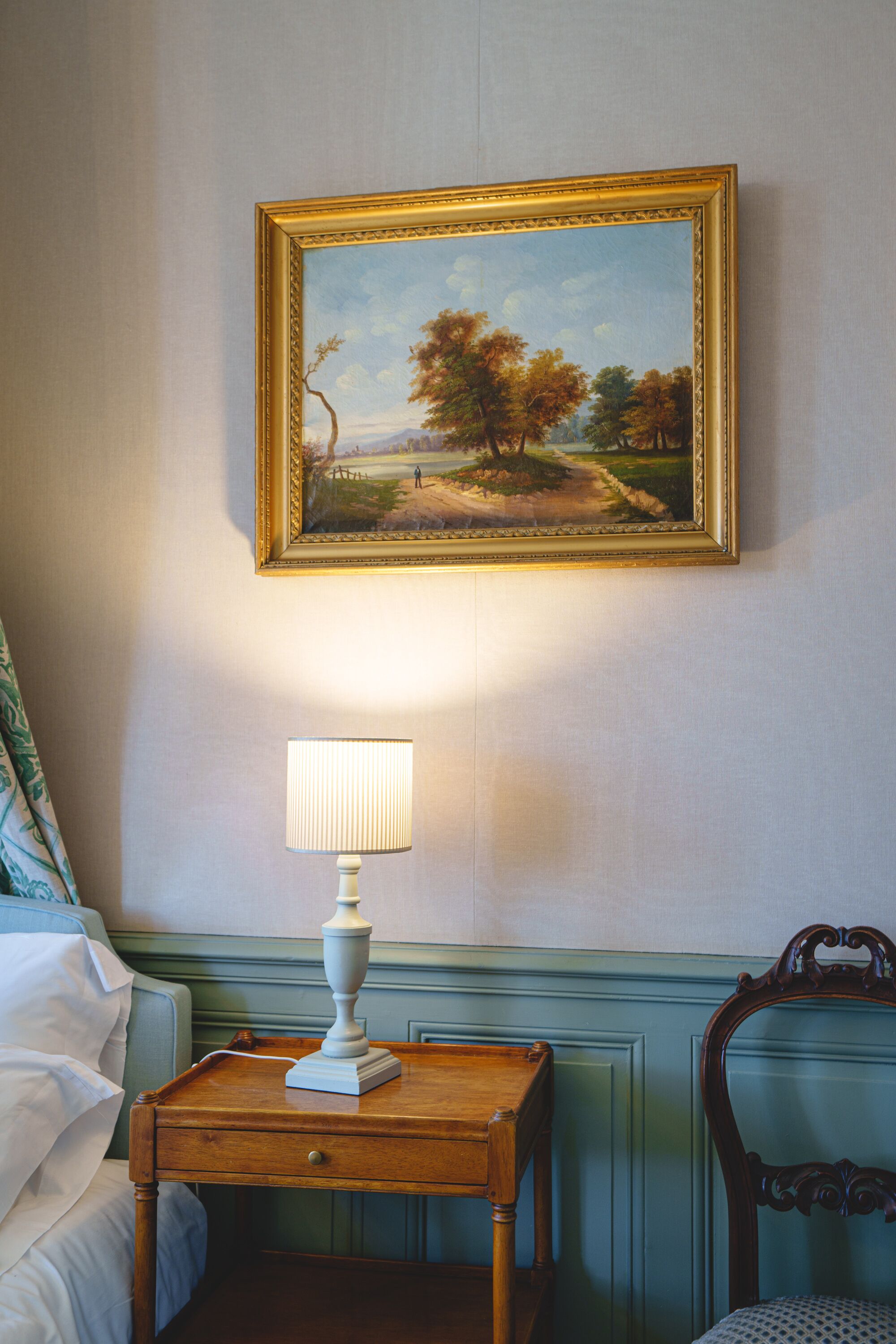 A bedroom with a bed, a nightstand with a lamp, a chair, and a framed painting on the wall.