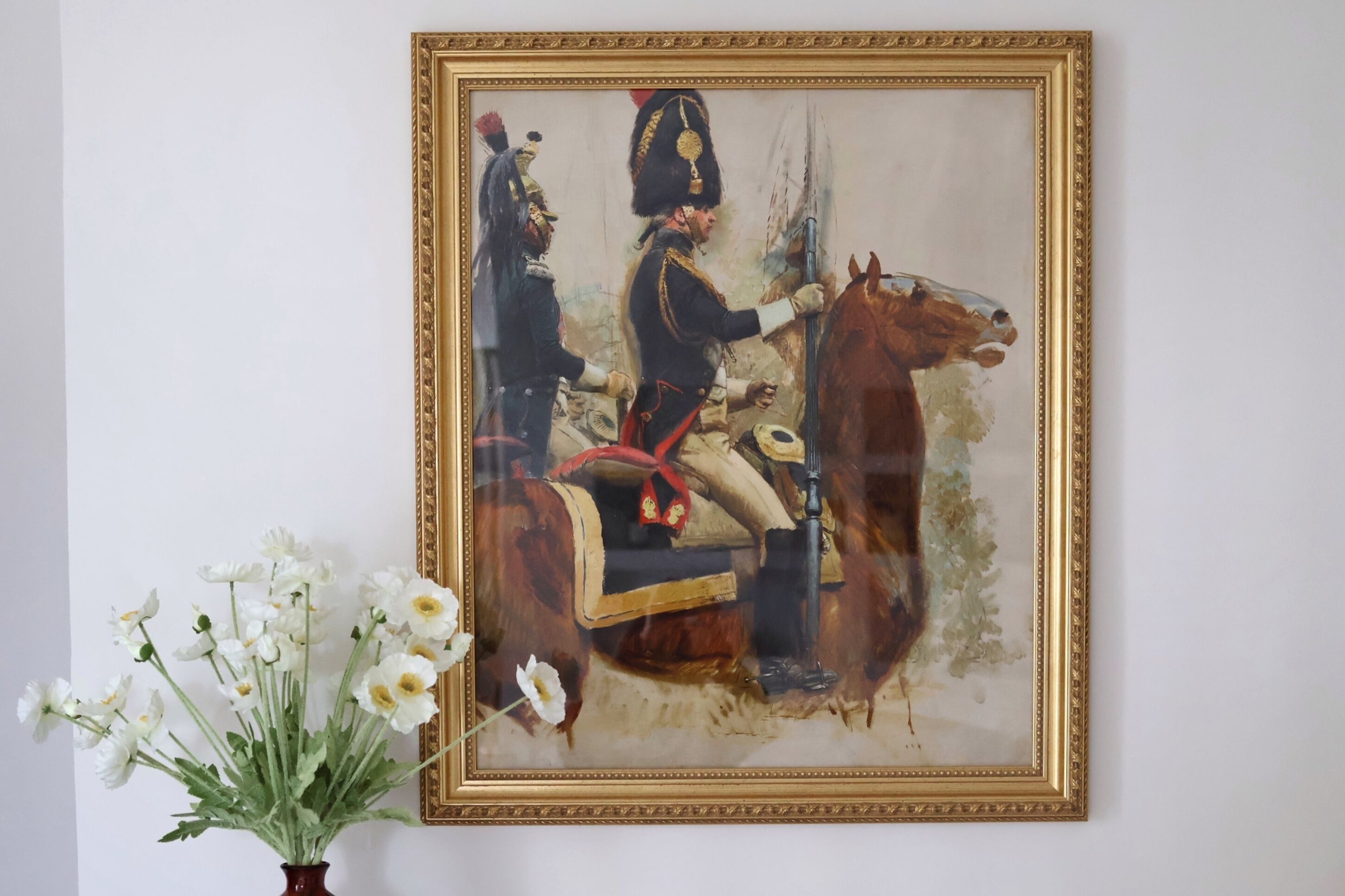 Un cuadro enmarco que muestra una figura montada con uniforme, un jarrón con flores blancas y un marco dorado.