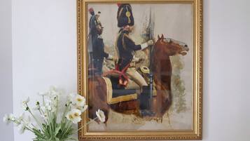 Un cuadro enmarco que muestra una figura montada con uniforme, un jarrón con flores blancas y un marco dorado.