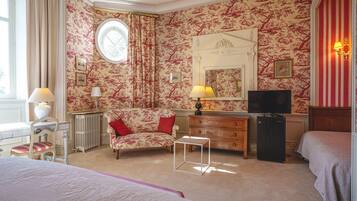 A hotel room with floral wallpaper, a round window, a bed, a dresser, a television, and a lamp.