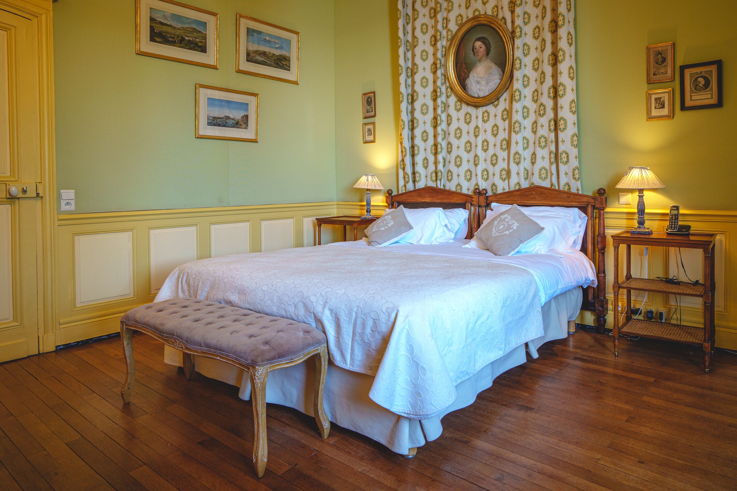 A bedroom with a large bed, a wooden bench, two bedside tables with lamps, and framed pictures on the walls.