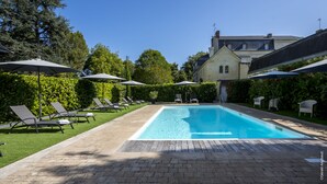 Una piscina rectangular con cubierta de madera, rodeada de sillas de descanso y sombrillas, con un edificio al fondo.